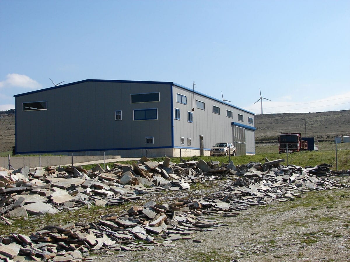 Final view of the steel building with panels and wind turbines in the background in Karystos